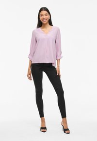 Blusa viola chiaro a collo a V con maniche a tre quarti arrotolate, abbinata a jeans skinny neri e tacchi aperti neri. Texture liscia.