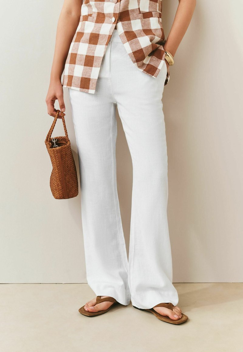 Personne portant un pantalon blanc à jambes larges, une chemise à carreaux marron et blanc, des sandales marron, tenant un sac à main marron tressé.