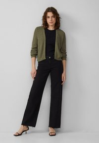 Cardigan vert olive avec tricot texturé, porté par-dessus un haut bleu marine avec de petites embellissements, associé à un jean noir à jambes larges et des sandales noires.