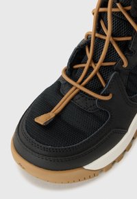 Gros plan sur une chaussure de sport noire avec un tissu en maille, des lacets marron et une semelle en caoutchouc, mettant en avant les détails de design et de texture.
