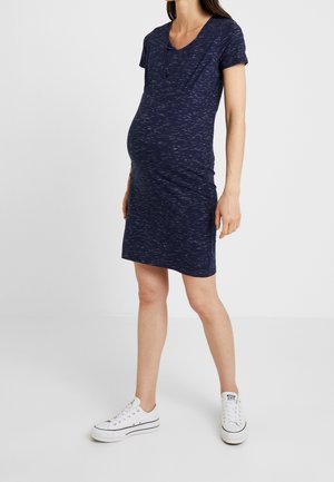 Gravid kvinde iført en tætsiddende marineblå kortærmet kjole og hvide platform sneakers, stående mod en ensfarvet lys baggrund.