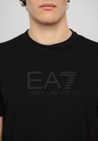 Melna kokvilnas T-krekls ar izceltu, tonālu „EA7” un „EMPORIO ARMANI” logotipu priekšpusē. Īsas piedurknes un apaļas kakla dizains.