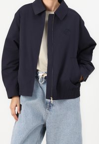 Marineblauw jack met een kraag, ritssluiting en elastische manchetten. Voorzien van een subtiel logo en zijzakken, gecombineerd met lichtblauwe jeans.