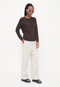 Pull en tricot marron, pantalon en velours côtelé crème et chaussures à plateforme noires avec une touche de rose. Le mannequin porte des lunettes de soleil surdimensionnées et se tient les mains dans les poches.