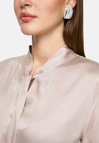 Silk blouse in light pink with a narrow collar and tie detail. The model wears large, shiny silver earrings. Smooth texture with subtle sheen.