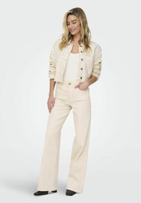 Veste en denim crème avec des détails en œillets, associée à un pantalon large beige clair et des mules à talons noires. Boutons en forme de rectangle sur la veste.