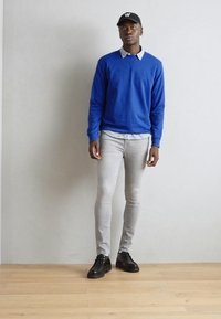 Suéter azul sobre una camisa blanca con cuello, jeans skinny gris claro y zapatos negros. El modelo está de pie contra una pared de color claro y un suelo de madera.