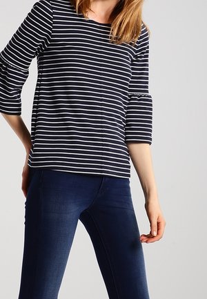 Kobieta w czarno-białej bluzce w poziome paski z rękawami do połowy przedramienia oraz w dopasowanych, ciemnoniebieskich dżinsach na jednolitym tle.