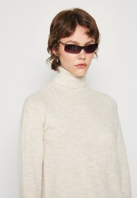 Petites lunettes de soleil rectangulaires avec des verres sombres, monture noire, et un pull col roulé ajusté en crème en tissu doux et texturé.