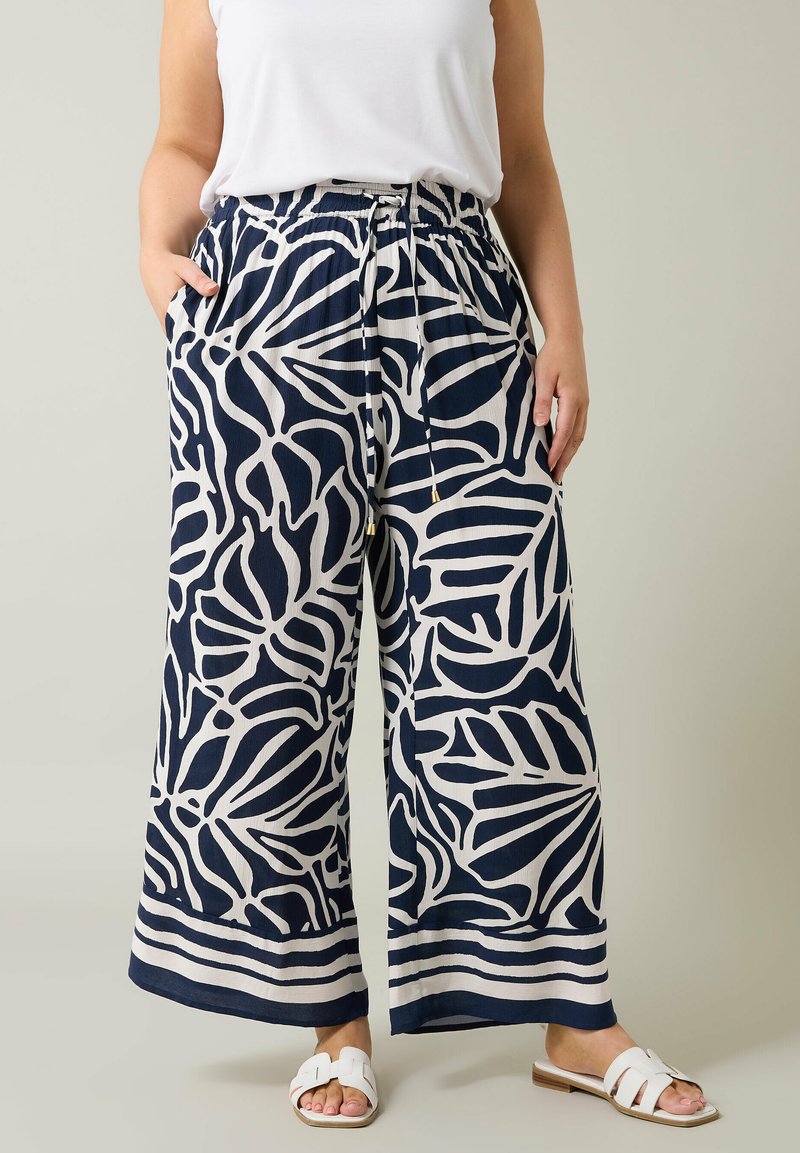 Pantalones de pierna ancha en azul marino y crema con un patrón de hojas, cintura elástica, cierre con cordón y bolsillos laterales. Combinados con una blusa blanca y sandalias.