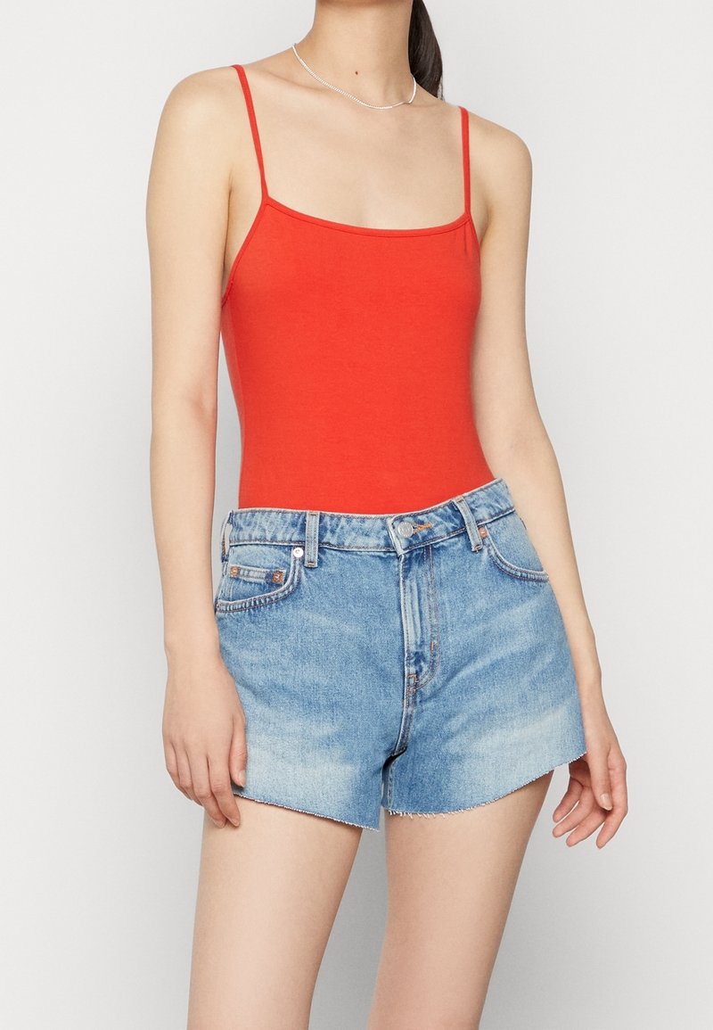 Röd ärmlös bodysuit i kombination med ljusblå denimshorts. Bodysuiten har en fyrkantig halsringning; shortsen har fransar i nederkant och fem fickor.