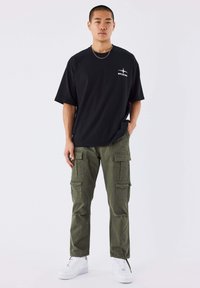 Jonge man die een zwart oversized T-shirt, olijfgroene cargobroek, witte sneakers en zilveren sieraden draagt, staand tegen een effen witte achtergrond.