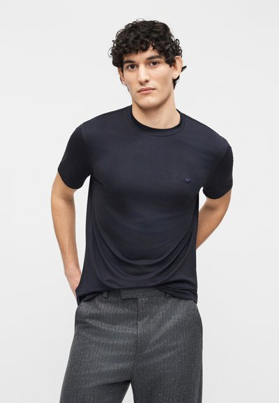 Νεαρός άνδρας με κοντά σγουρά μαλλιά, φορώντας εφαρμοστό μαύρο κοντομάνικο πουκάμισο και γκρι υφασμάτινο παντελόνι, στέκεται μπροστά σε απλό φόντο.