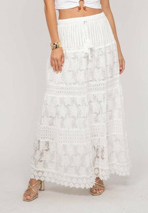 Femme portant une longue jupe blanche en dentelle avec des motifs floraux, une taille crochetée et des liens à pompons, associée à des talons dorés à brides et un bracelet doré.