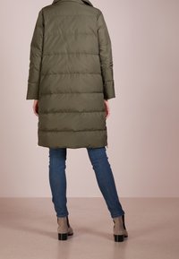 Olivgrüner Puffer-Mantel mit horizontalen Nähten, langen Ärmeln und mittellanger Schnittführung. Kombiniert mit blauen Jeans und braunen Knöchelstiefeln.