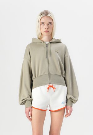 Haut à capuche olive ouvert avec une fermeture éclair, présentant des manches larges. Associé à un short blanc avec des accents orange au niveau de l'ourlet et du cordon de serrage.