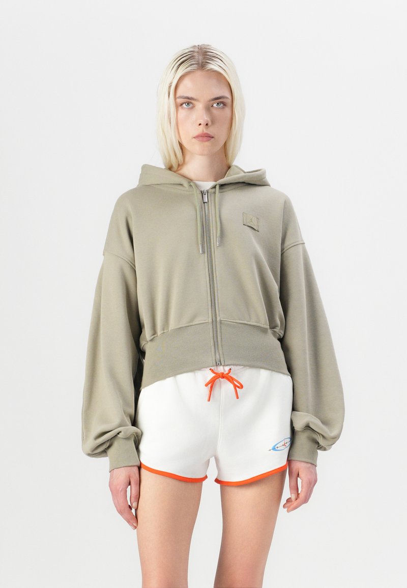 Sudadera verde oliva recortada con cremallera y capucha, que cuenta con mangas amplias. Combinada con pantalones cortos blancos con detalles en naranja en el dobladillo y el cordón.