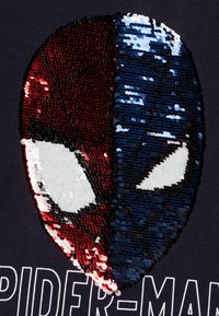 Design eines Spider-Man-Maskenmotivs in Rot und Blau aus Pailletten auf einem dunklen T-Shirt; mit strukturierten Pailletten, kontrastierenden Farben und großen weißen Augenakzenten.
