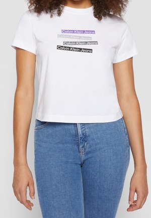 Femme portant un crop top blanc à manches courtes avec le texte "Calvin Klein Jeans" en quatre barres colorées et un jean bleu taille haute.