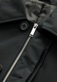 Chaqueta negra con un cierre plateado, un destacado detalle de botones y una textura mate suave con un forro acolchado.