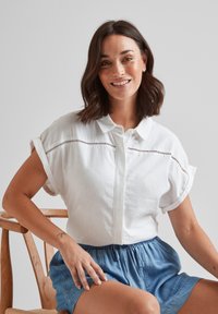 White short-sleeve shirt with a button front and eyelet detailing. Paired with light blue drawstring shorts. Wood chair in background.
