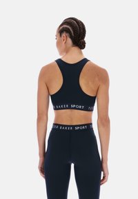 Moteris su pintais plaukais, vilkinti juodą sportinį liemenėlę ir tamprėmis su "TED BAKER SPORT" ženklu, rodoma iš nugaros ant baltos fono.