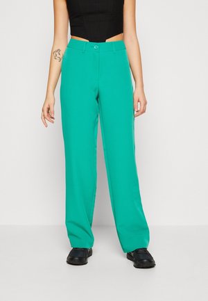 Vrouw met turquoise rechte broek, zwart crop top en zwarte schoenen, staand tegen een effen lichte achtergrond.