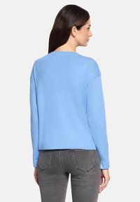 Jasnoniebieski sweter z długimi rękawami, okrągłym dekoltem oraz lekko skróconym dołem, wykonany z miękkiego materiału. Noszony z szarymi dżinsami, o minimalnym wzornictwie.