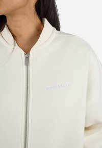 Cremfarbener Bomberjacke mit durchgehendem Reißverschluss, geripptem Kragen und dezent gesticktem Logo auf der Brust. Weicher, strukturierter Stoff.