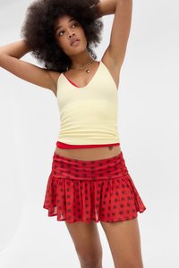 Młoda kobieta z naturalnymi afro włosami ma na sobie żółty top na ramiączkach na czerwonym biustonoszu i czerwoną spódniczkę w czerwone kropki, pozując z uniesionymi rękami.