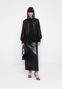 Blusa nera trasparente con colletto a fiocco, abbinata a una lunga gonna nera di pelle. La modella tiene in mano una borsa nera e indossa sandali con cinturini.