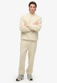 Giovane uomo in piedi con le braccia incrociate, indossa una felpa beige e pantaloni tuta, con sneakers grigie e bianche, su sfondo bianco.