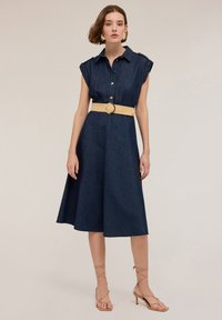 Denim-kangashirt dress with short sleeves, a button front, and a flared knee-length skirt, paired with a tan woven belt and strappy beige heels.