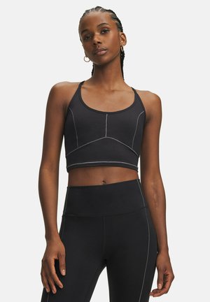 Naine, kellel on punutud juuksed, musta spordirinnahoidja ja kõrge vöökohaga retuusid, seisab lõdvestunud kehahoiakuga tavalise valge tausta ees.