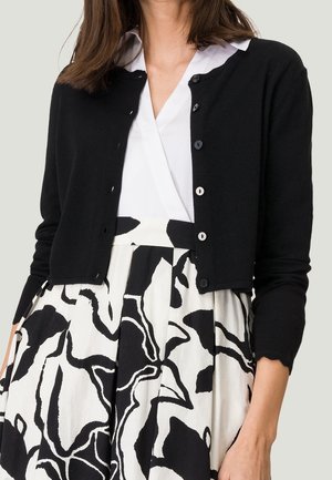 Cardigan noir à manches longues, col rond, et fermeture à boutons, porté sur une blouse blanche et une jupe à motif noir et blanc.