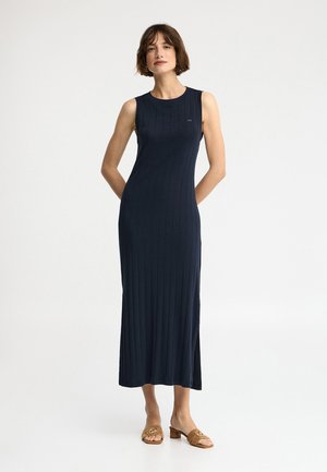 Frau steht mit den Händen hinter dem Rücken, trägt ein ärmelloses marineblaues geripptes Midikleid und hellbraune offene Sandaletten mit Absatz vor weißem Hintergrund.