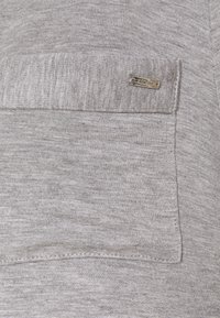 Γκρι βαμβακερό ύφασμα με υφή στην επιφάνεια, διαθέτει μια μικρή τσέπη στο στήθος και μεταλλικό λογότυπο. Απαλή, ελαφριά και casual σχεδίαση.