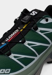 Πράσινο αθλητικό παπούτσι με υφαντή ανώτερη επιφάνεια, λευκές και μαύρες λεπτομέρειες, ελαστικό σύστημα κορδονιών και λογότυπο στη γλώσσα.