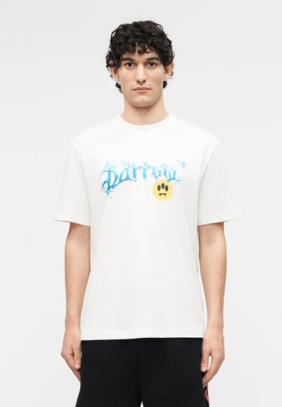 Νεαρός με σγουρά μαλλιά που φοράει λευκό t-shirt με μπλε γοτθικό κείμενο και κιτρινισμένο εικονίδιο απογοητευμένου συναισθήματος με δόντια.