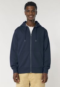 Sweat à capuche zippé bleu marine en tissu doux, avec une poche avant, des cordons de serrage et des poignets côtelés. Porté par-dessus une chemise claire.