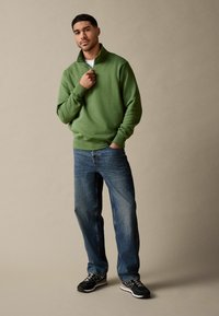 Green zip-neck sweatshirt with long sleeves, paired with blue jeans and black sneakers. Set against a neutral background.