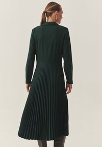 Vestido plisado de un verde oscuro, con mangas largas y un cuello con solapa, que presenta un cuerpo ajustado y una falda fluida. Textura de tejido suave.