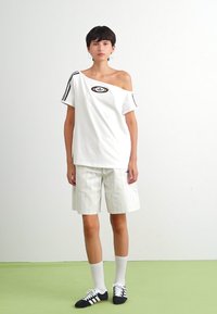 Balta off-the-shoulder t-kreklu ar melnu logo, kombinē ar baltām svītrainām šortiem. Modelis valkā melnas kurpes un baltas zeķes.