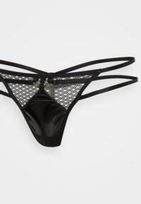 Musta thongi, joka on valmistettu verkkomaisesta ja kiiltävästä materiaalista, jossa on useita hihnoja ja metallikoristeita. Suunniteltu minimalistiseksi, avoin selkäpuoli.