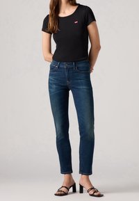 T-shirt nera a maniche corte con un piccolo logo rosso, abbinata a jeans scuri aderenti e sandali neri con tacchi e fasce sottili.