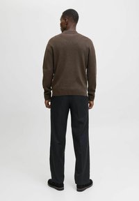 Muž s krátkými vlasy a náušnicí, otočený zády, oblečený v hnědém svetru s dlouhým rukávem, černých kalhotách a černých botách, na jednolitém pozadí.