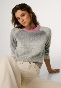 Suéter de punto gris con un cuello acanalado rosa, que presenta un corte holgado y mangas raglán. El modelo lleva pantalones beige, sentado contra un fondo claro.