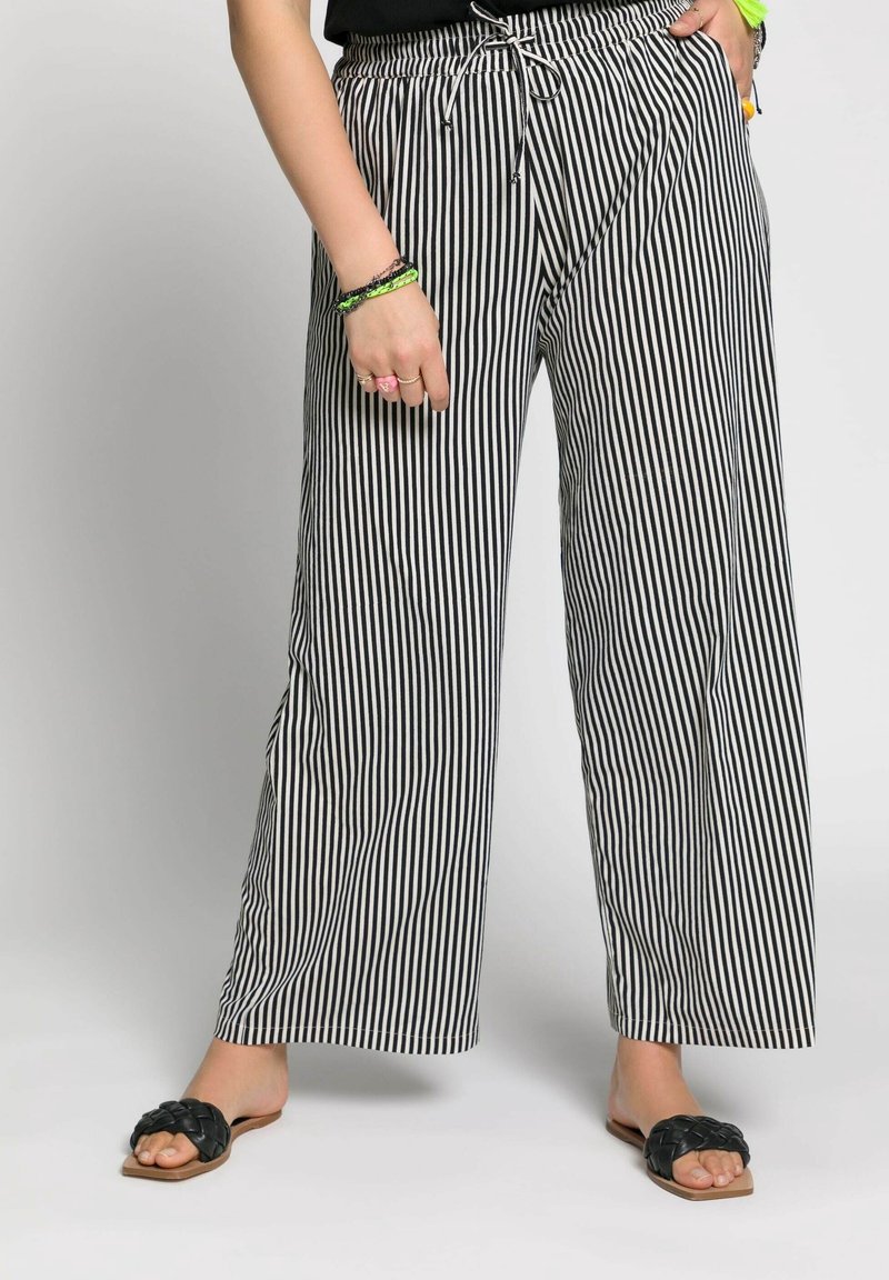 Femme portant un pantalon large à rayures verticales noires et blanches avec des sandales noires tressées sans lacets, debout les mains dans les poches.