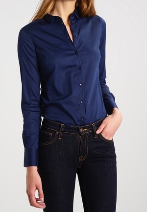 Kobieta ubrana w dopasowaną granatową koszulę na długi rękaw z guzikami, wpuszczoną w ciemnoniebieskie dżinsy z pomarańczowym przeszyciem, ręka spoczywa w kieszeni.