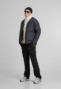 Polstret sort bomberjakke med quiltet tekstur, tætsiddende hvid turtleneck, sorte cargo bukser og reflekterende sneakers, accessoireret med solbriller.